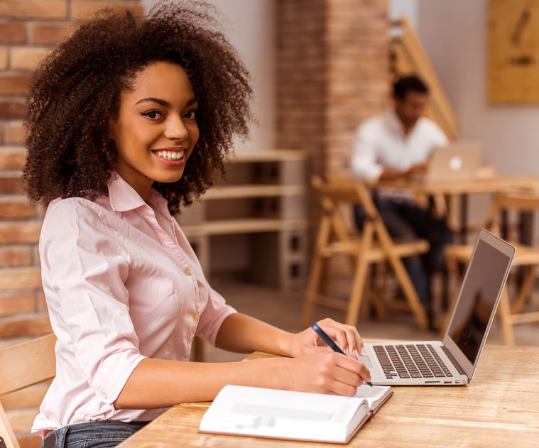 young beautiful afro-american woman looking in camera, smiling, using laptop and writing in notebook while studying in cafe concept for Scholarship Essay Tips