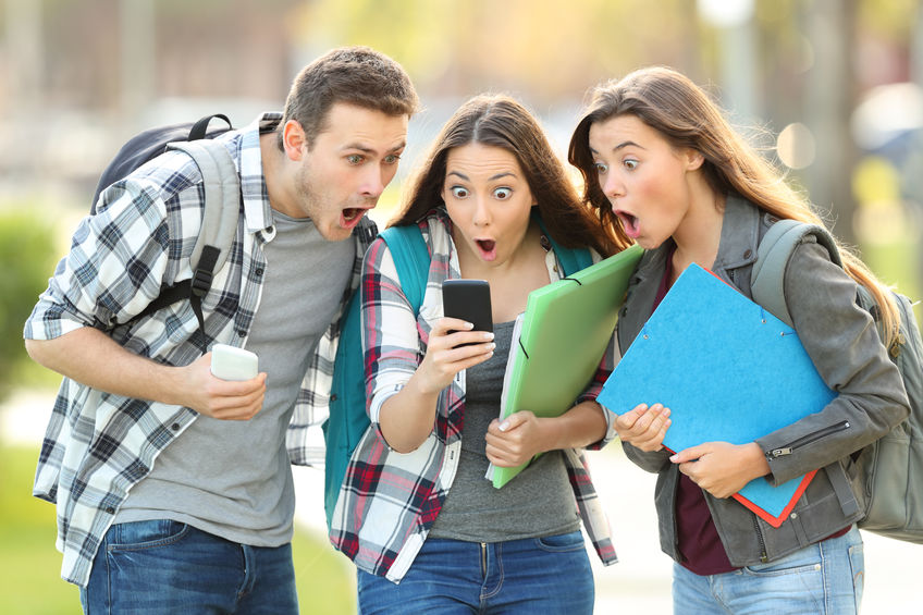 Three amazed students checking on line content on a smart phone in the stre...