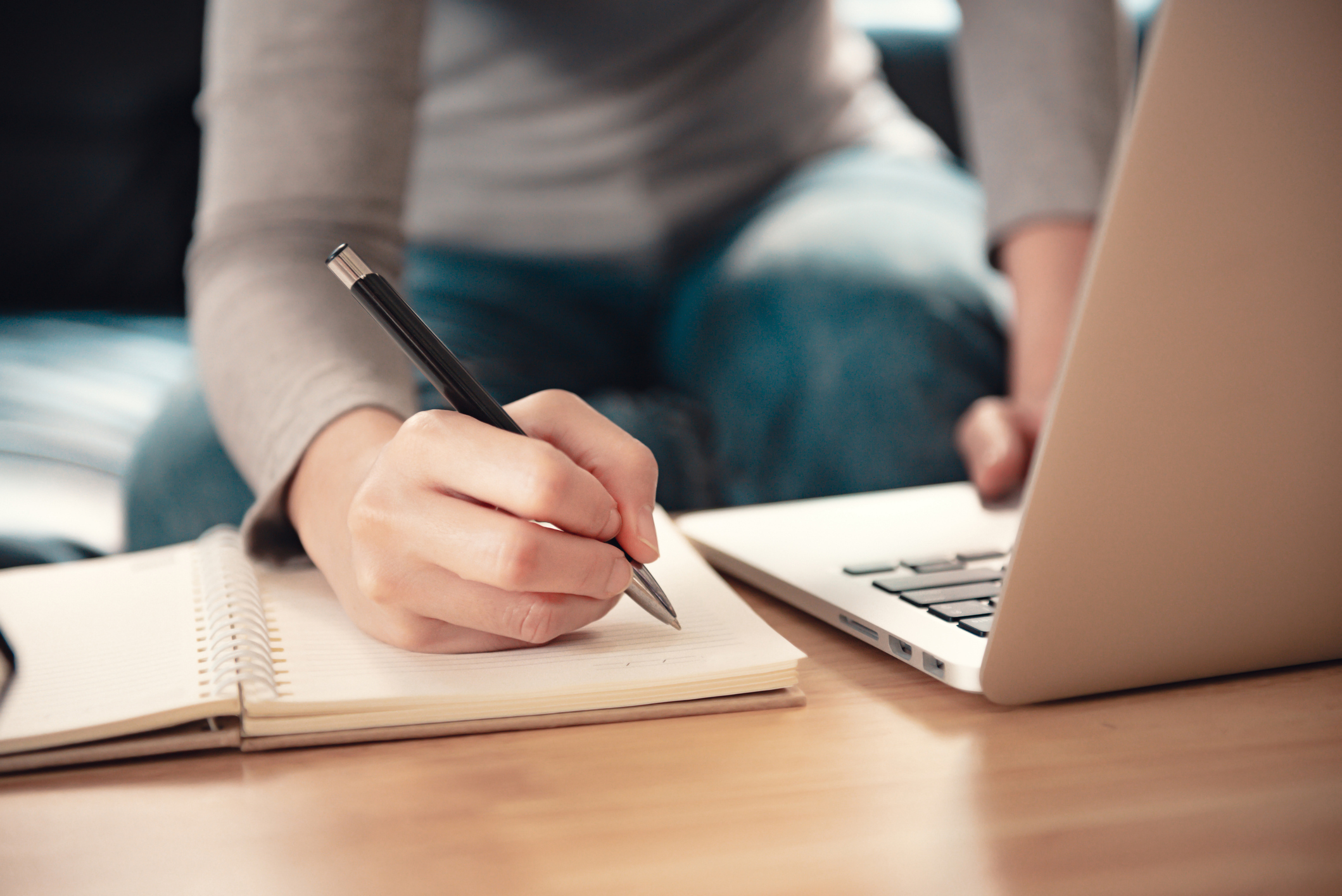 Woman writing on notebook and using laptop on sofa.