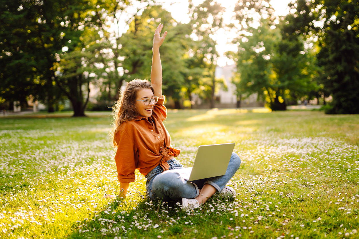 Young woman is sitting in a sunny park on a clearing and working with a laptop.