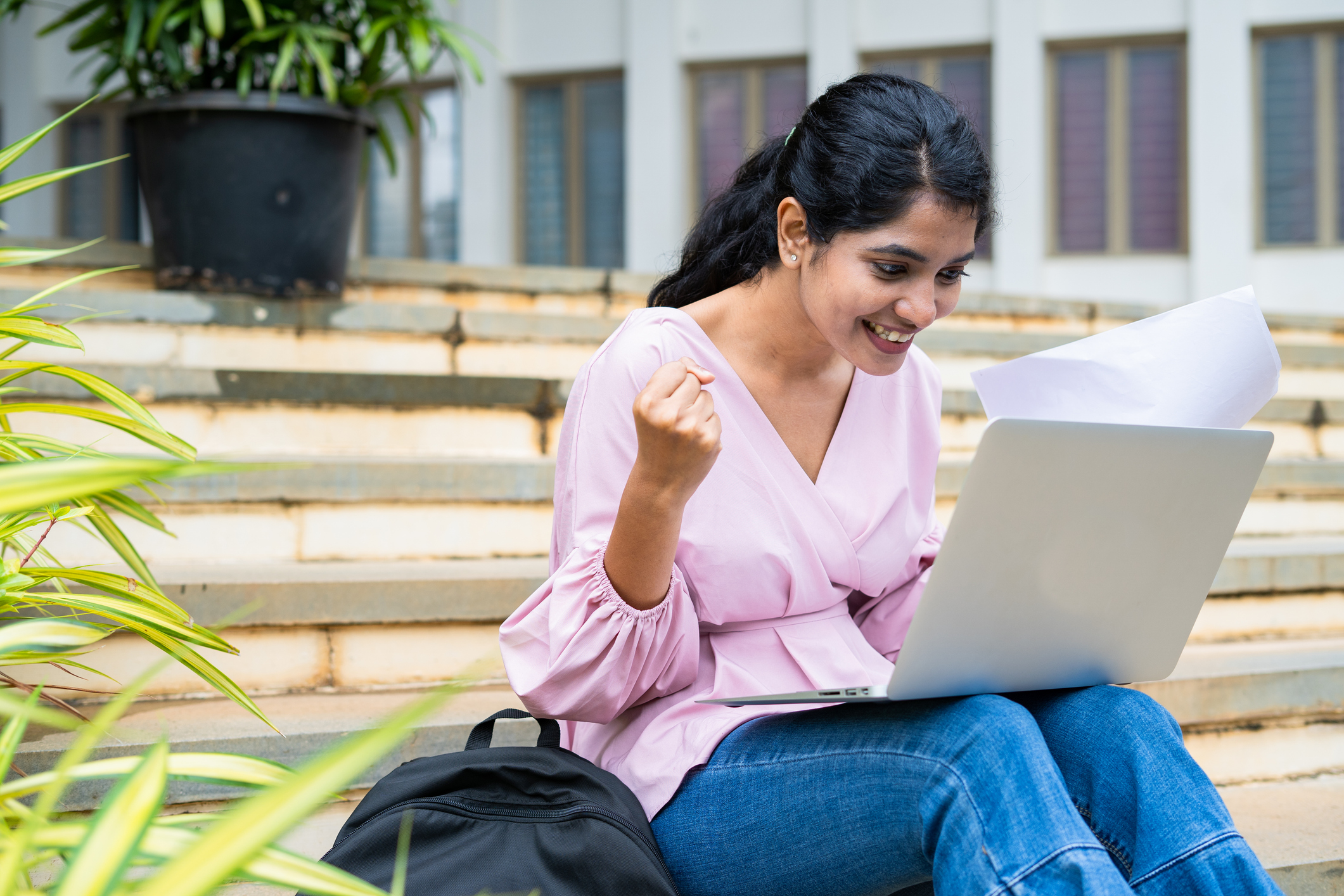 Excited girl students celebrating after seeing result on laptop while sitting on collage campus - concept of achievement, education and technology