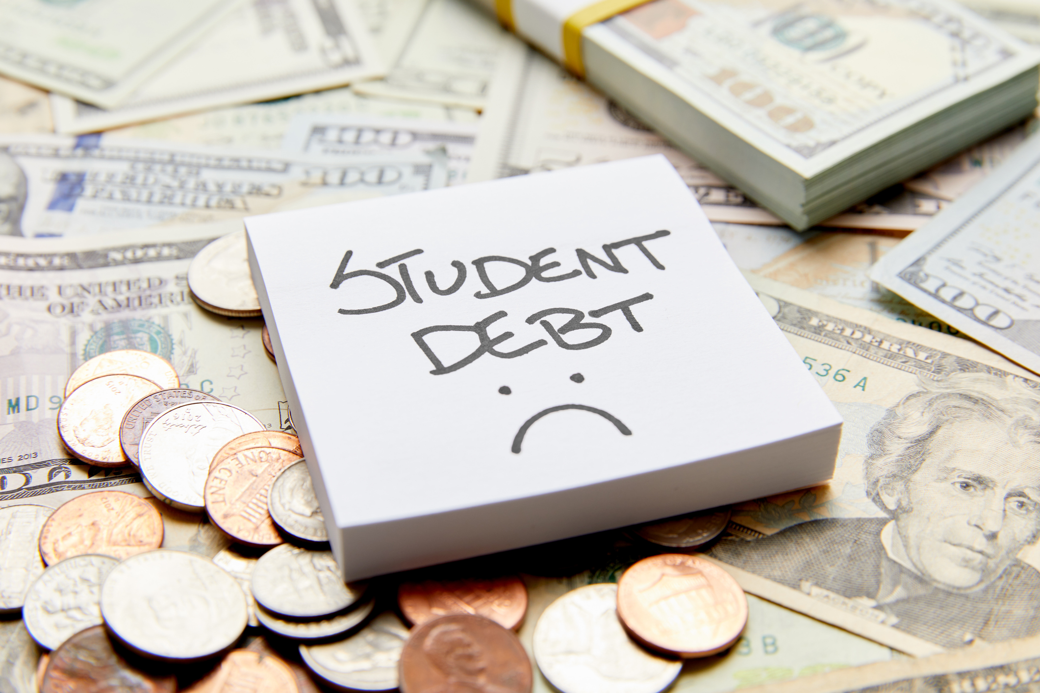 Student debt with sad face written on white sticky note on top of cash money with stack of money and coins