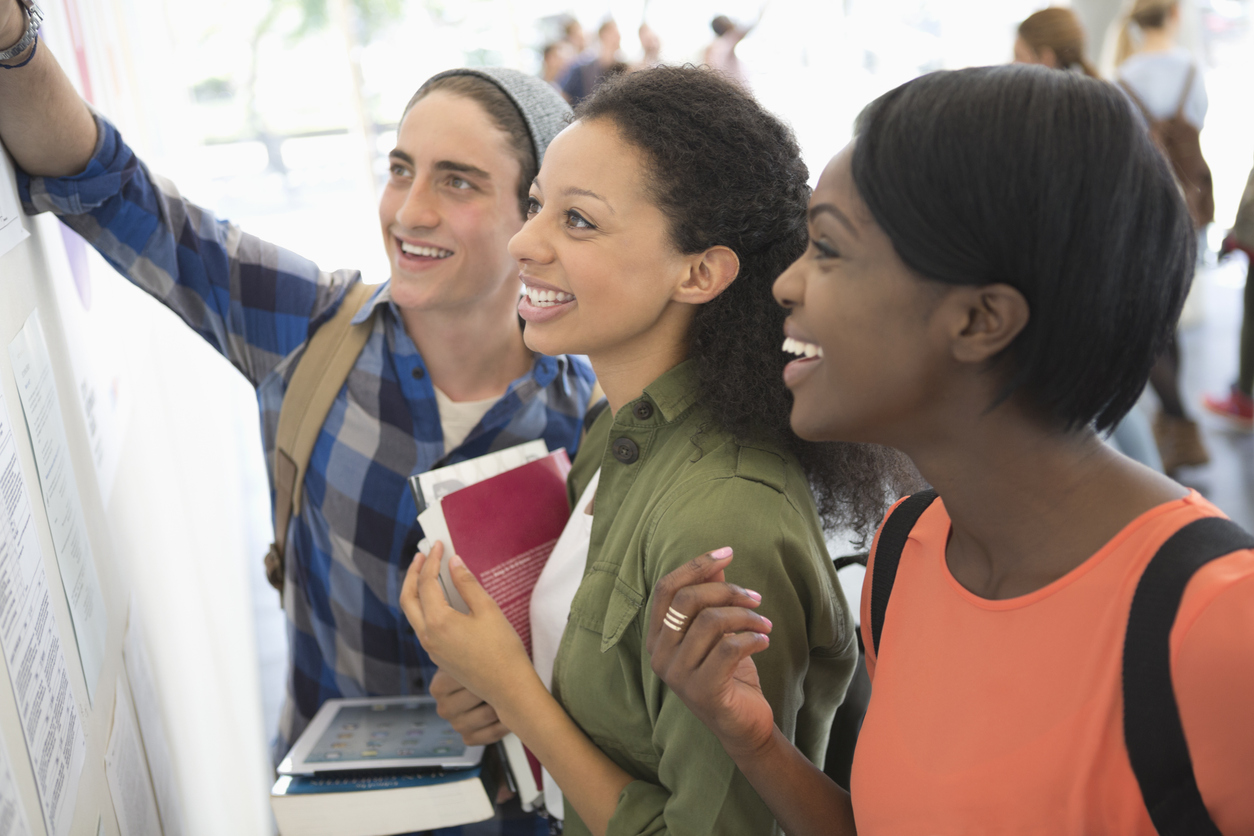 Three smiling students looking at information board, holding books and digital tablet 